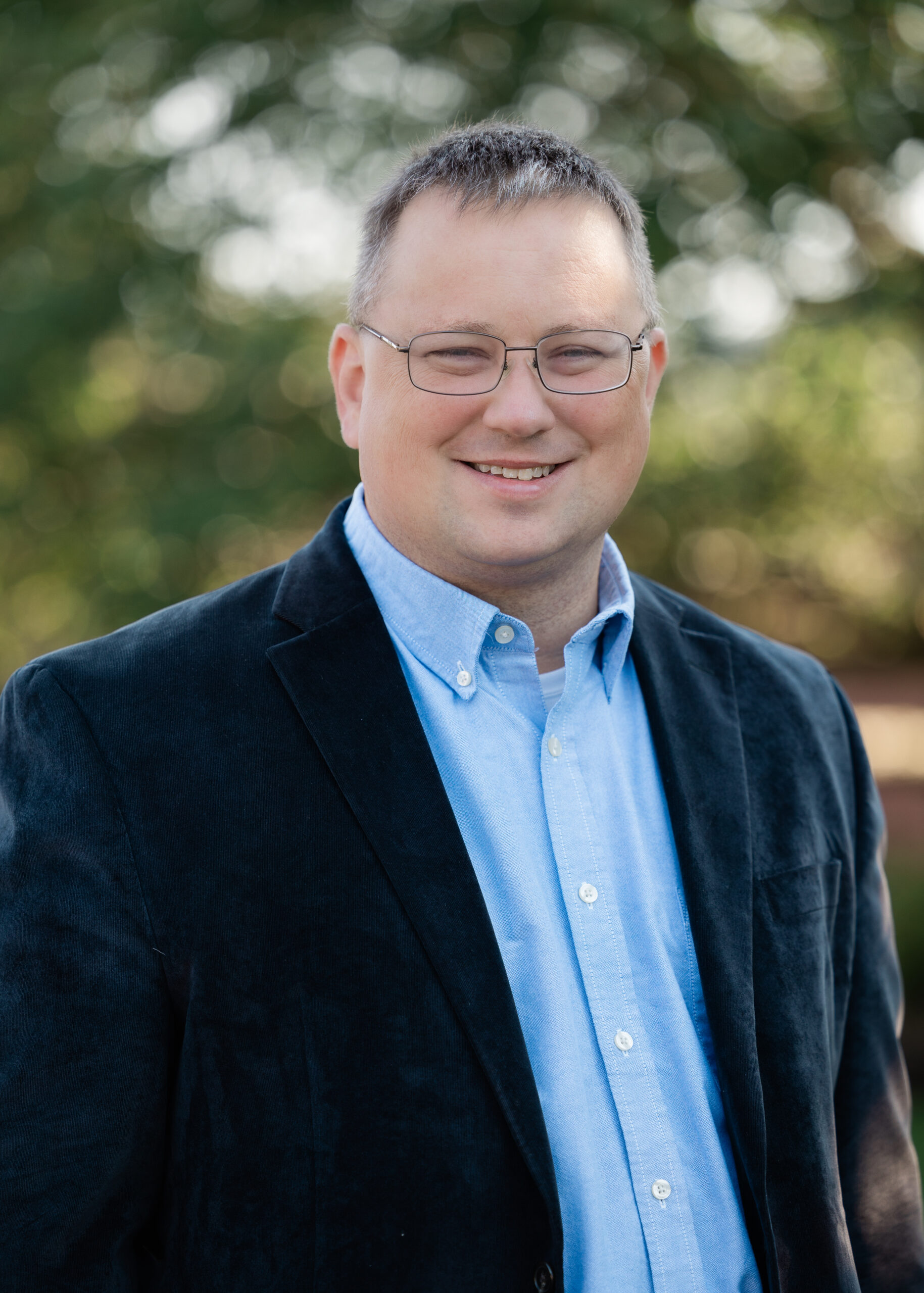 White male with glasses and a dark blue sport coat and light blue button up shirt in from of a blurry landscape background.
