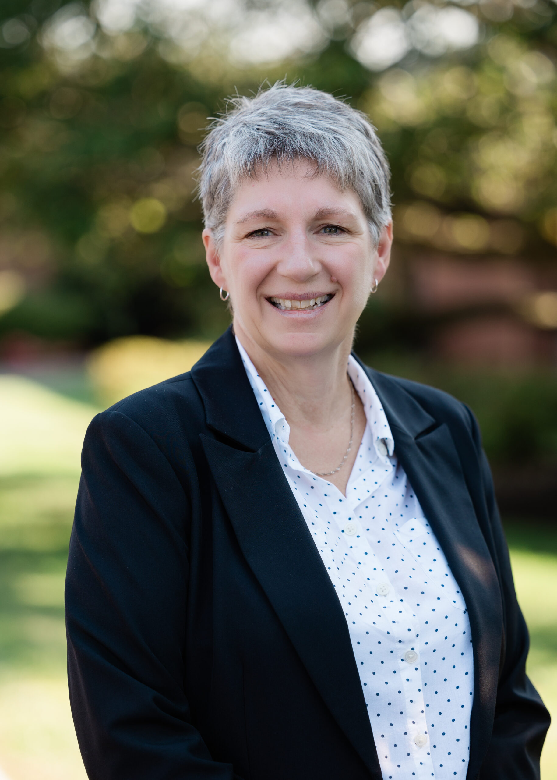 White woman with short gray hair wearing a black blazer and white button up shirt with black spots.